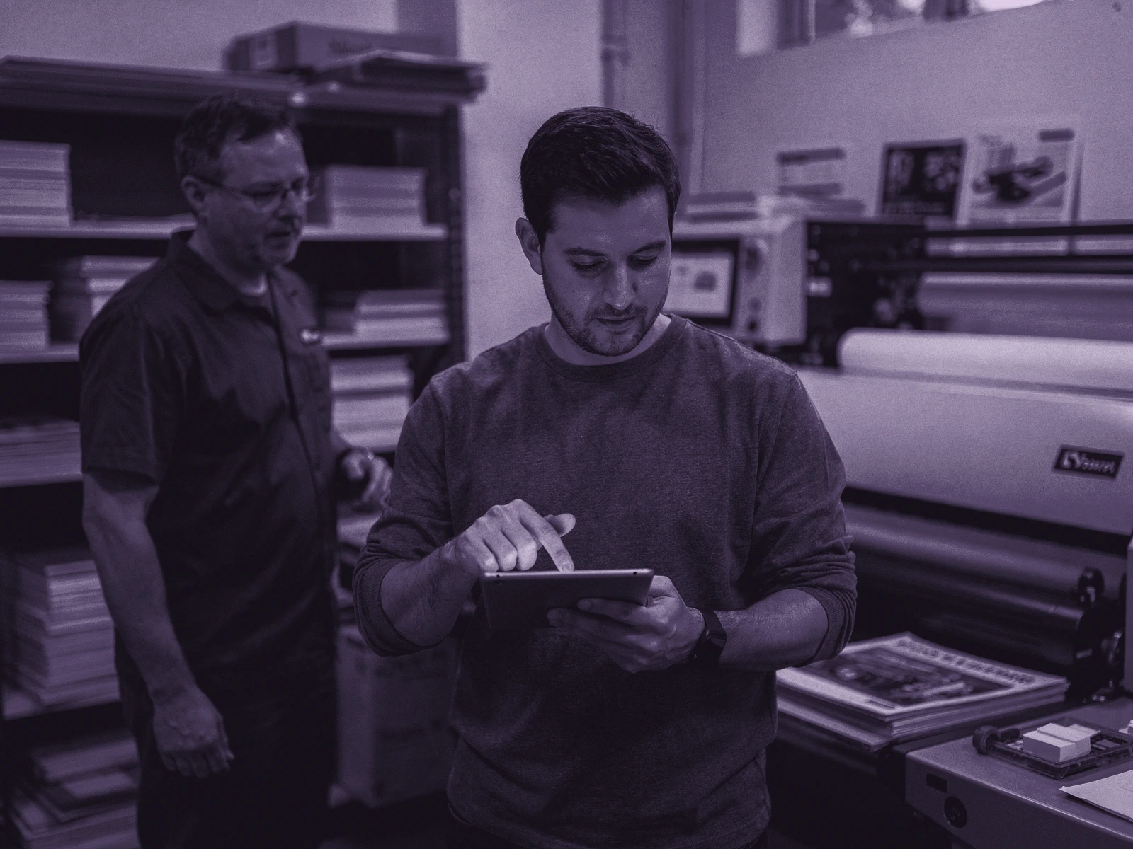 A project lead carefully reviews a digital proof on a tablet before a colleague proceeds with production